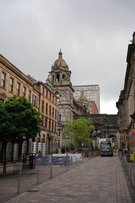 A wide urban street scene in Glasgow showing the historic architecture of Buchanan Street, with a prominent stone building featuring a domed clock tower and ornate details, flanked by modern office bu