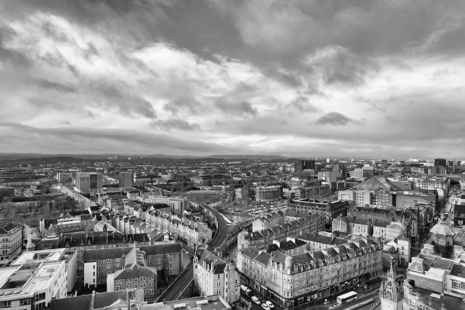 A black and white aerial photograph of Glasgow's cityscape taken during daytime, showing a dense arrangement of multi-storey and terraced buildings, roads, and bridges. Visible are various residential